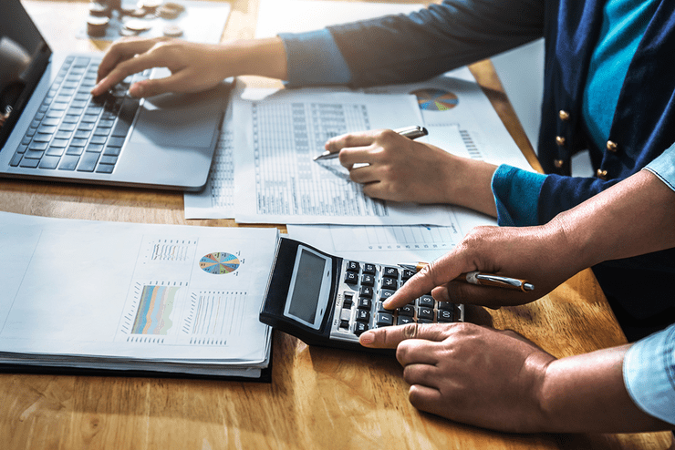 Ilustračná fotografia - ruka pracuje na notebooku a vytlačených tabuľkách, druhá osoba počíta na kalkulačke