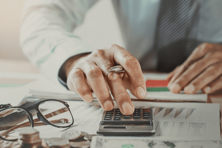 Ilustračná fotografia - ruka s perom nad kalkulačkou, na stole položené grafy, okuliare a mince